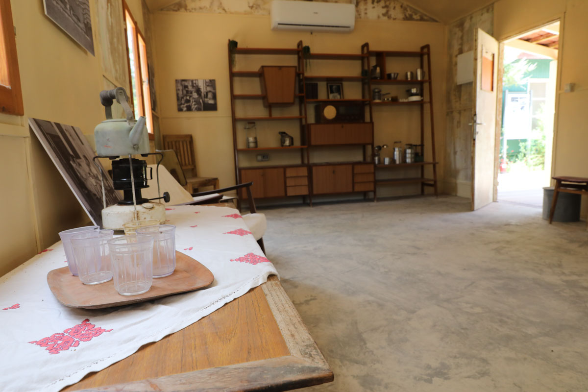 מבט מתוך צריף הראשונים, בקידמת התמונה נראה שולחן ועליו כלים ישננים וברקע שידת מדפים ענקית של פעם ותמונות בחור לבן.