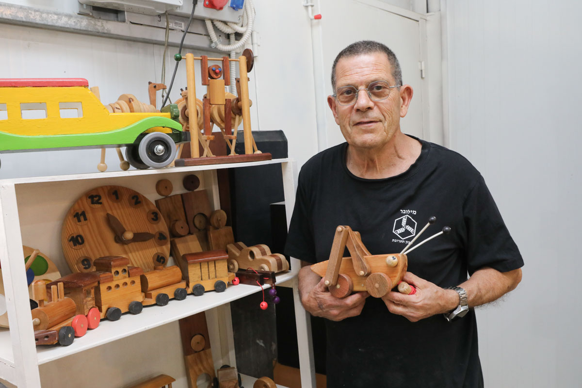 שי קורן עומד לצד מדפי הצעצועים שיצר ואוחז צעצוע חרגול עץ.