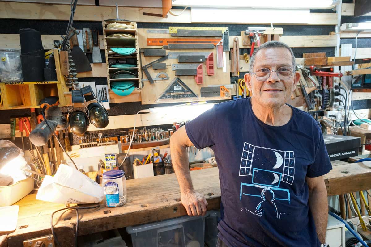 שי קורן עומד לצד שולחן עבודה ועל הקיר המון כלים מסוגים שונים להם הכין מקום במיוחד על קירות הנגריה שלו בשלל פטנטים.