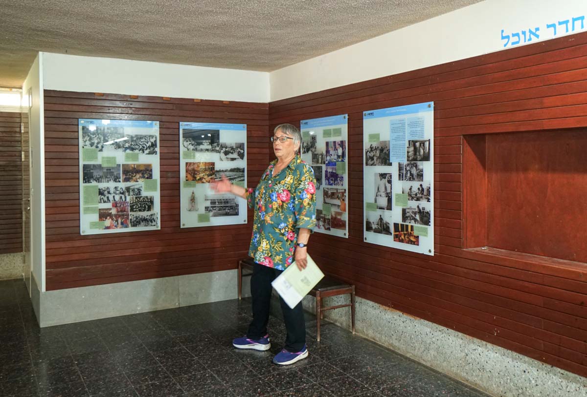 טלי שנר, יוצרת הסיור המודרך בקיבוץ, עומד לפני חלק מתערוכת "סיורי חדר אוכל"- שלטית המציגים סיפורים ואירועים מחדר האוכל בקיבוץ.