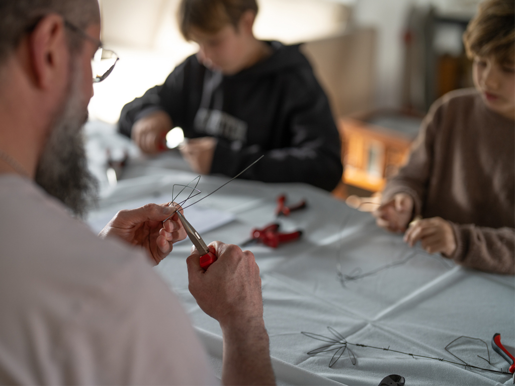 צילום מעבר לכתפו של האומן קובי סיבוני בזמן שהוא מכין יצירה ומעביר סדנא לשני ילדים בעבודה בחוטי הברזל.