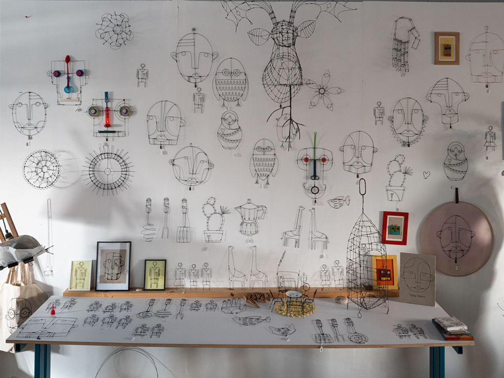 תמונה של קיר בסטודיו המציג את יצירותיו של קובי סיבוני, לפני הקיר מוצג שולחן תצוגה ועליו יצירות נוספות. רובן יצירות דו-מיימדיות המדמות פרצופים, חפצים, אנשים, נופים, צמחים וחיות.